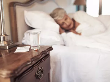 Medicijnen en een glas water met op de achtergrond een oudere vrouw slapend in haar bed